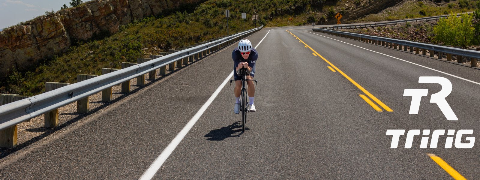 A cyclist on an open highway with guardrails, rugged hillside to the left, and a "TR TRiRiG" logo in the corner.